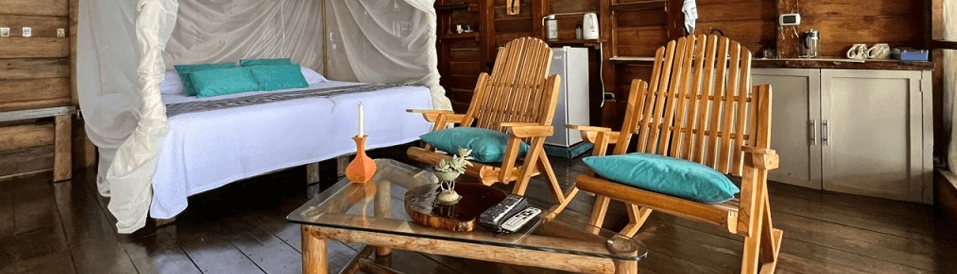 A cozy wooden room featuring a bed with a mosquito net, two wooden chairs, and a glass coffee table.