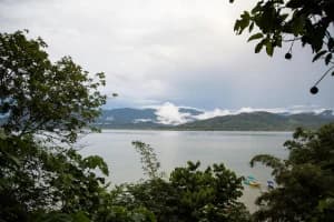A cloudy view of a calm lake surrounded by lush greenery and distant mountains.