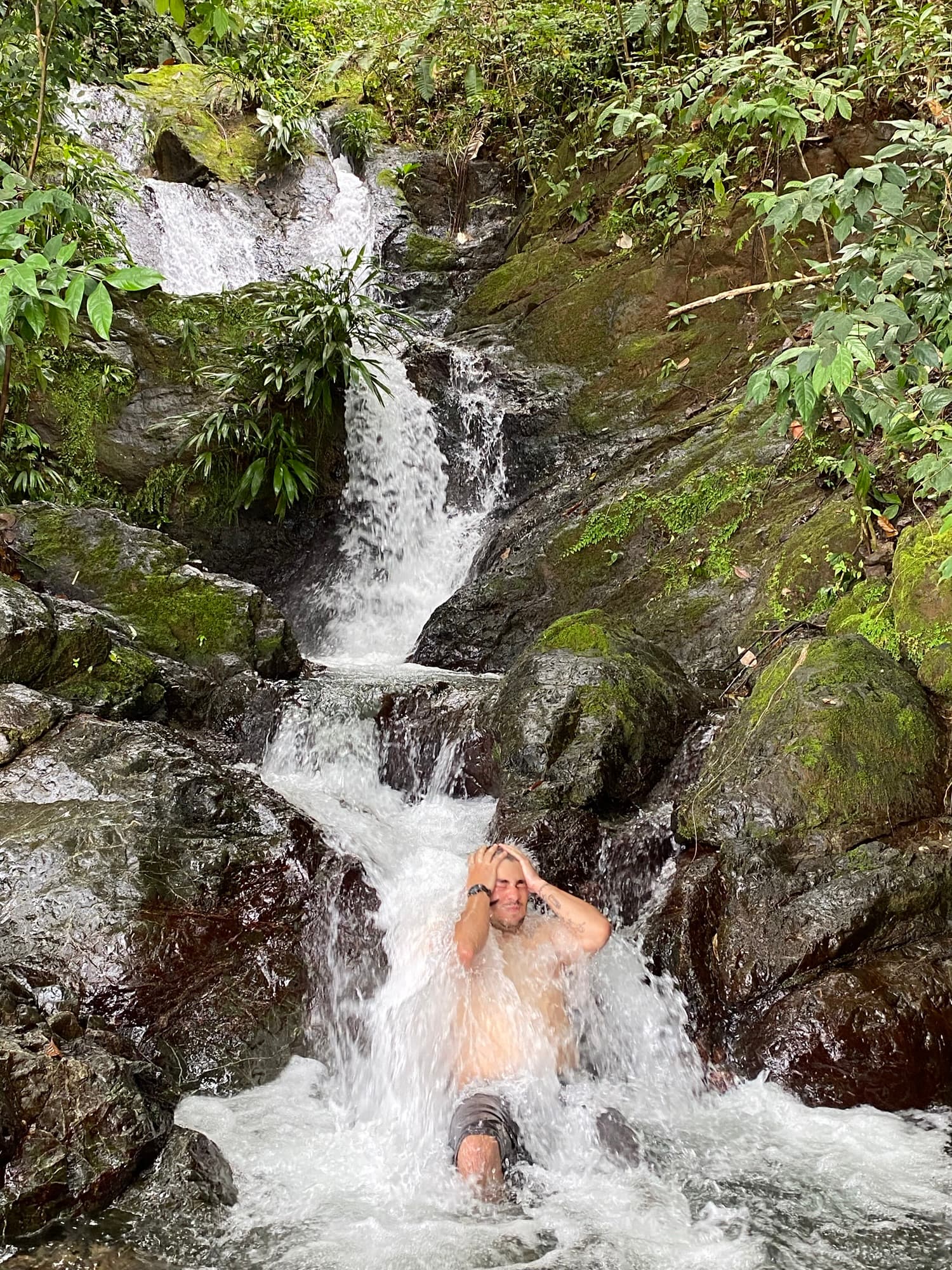 A man sits in a waterfall surrounded by lush greenery, water splashing around him.