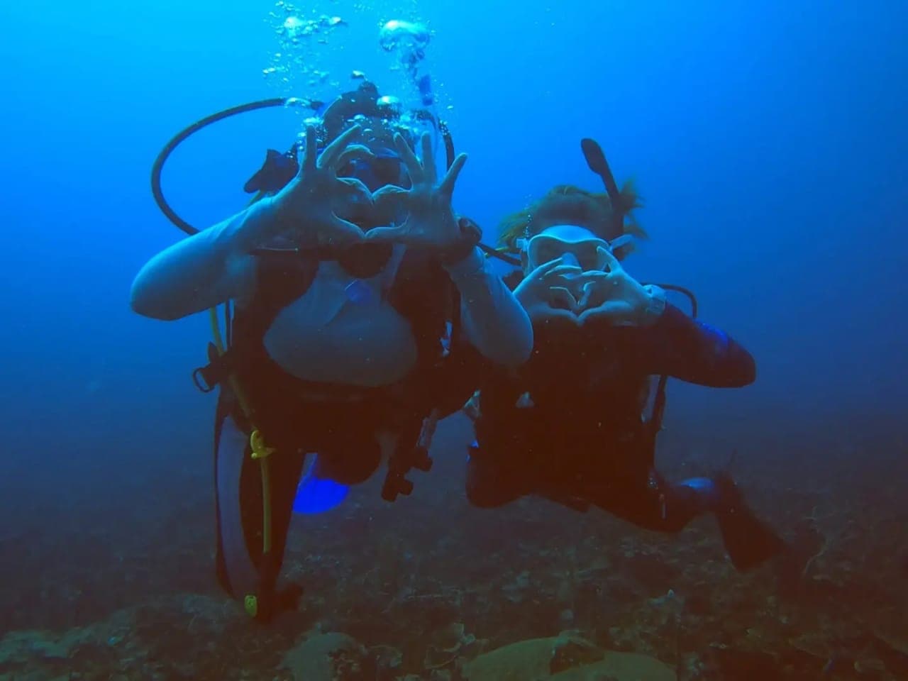 Two divers in scuba gear making heart shapes with their hands underwater.