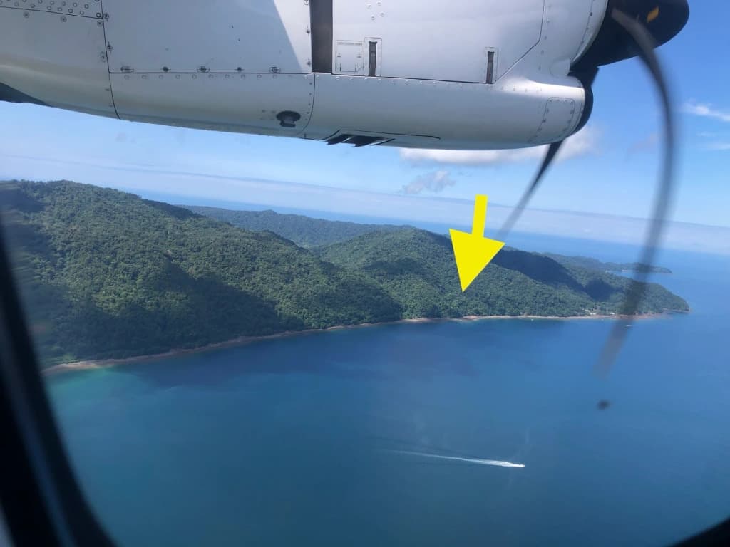 Aerial view of a coastline with lush green hills and a visible boat on the water, with the airplane's wing in the foreground.