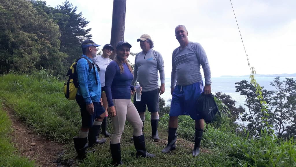 A group of five people dressed for hiking stands on a green trail with a scenic view of the ocean in the background.