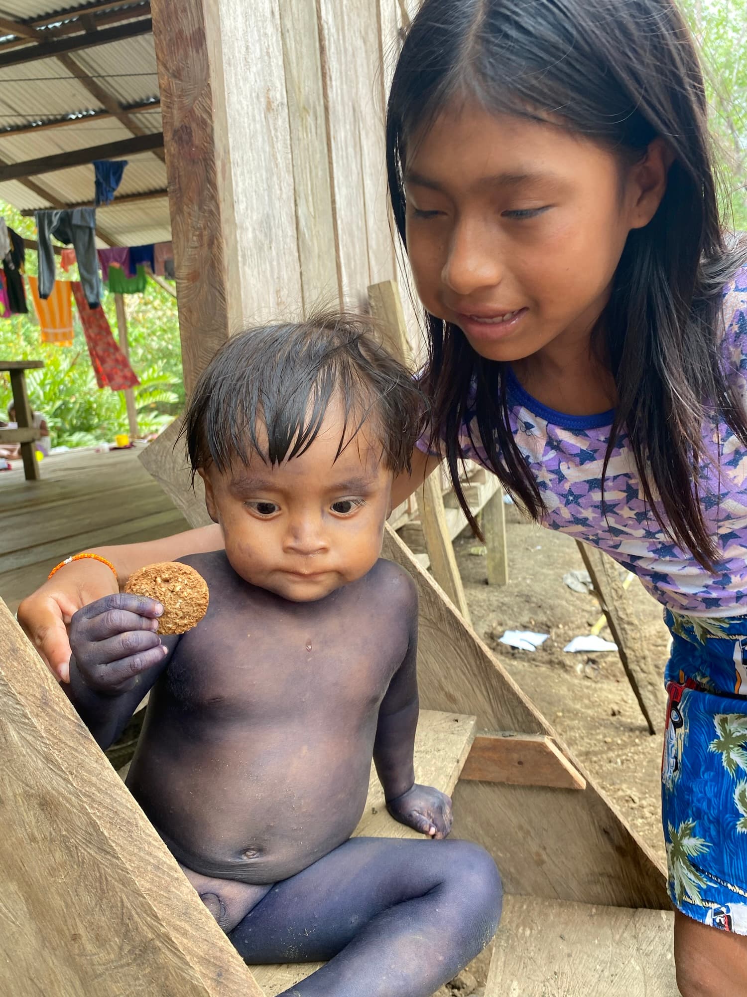 A young girl smiles at a baby sitting on wooden steps, holding a cookie while both appear dirty from play.