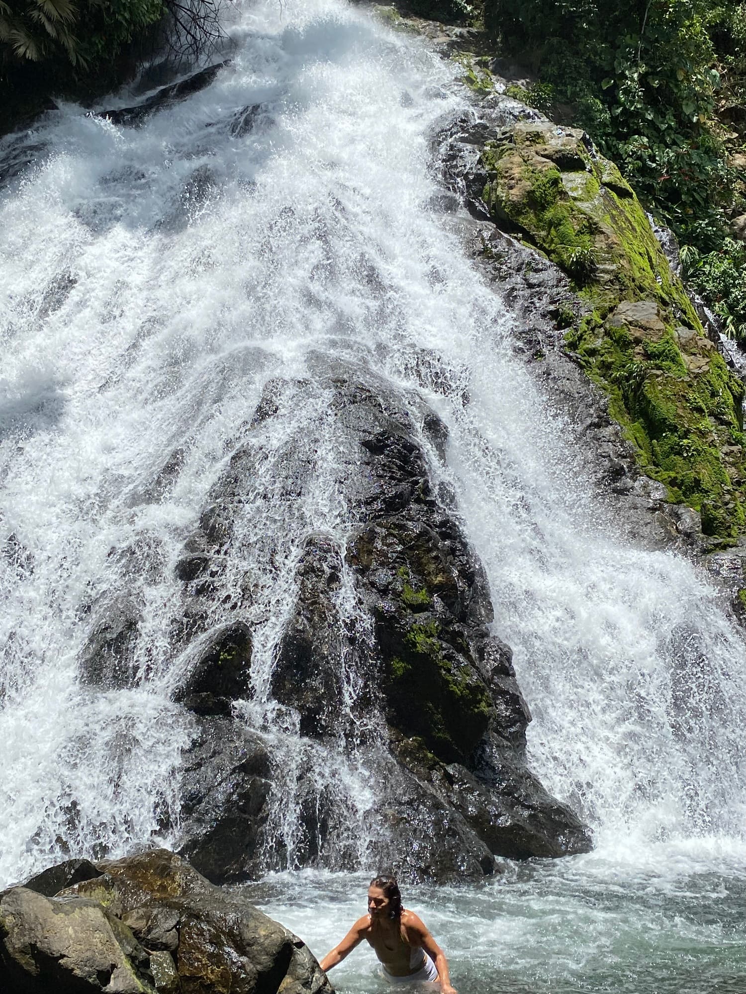 A woman wades in a natural pool at the base of a cascading waterfall surrounded by lush greenery.