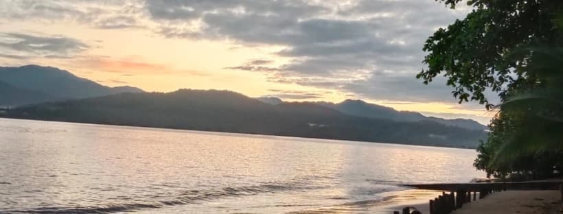 A serene beach at sunset, with mountains in the background under a cloudy sky.