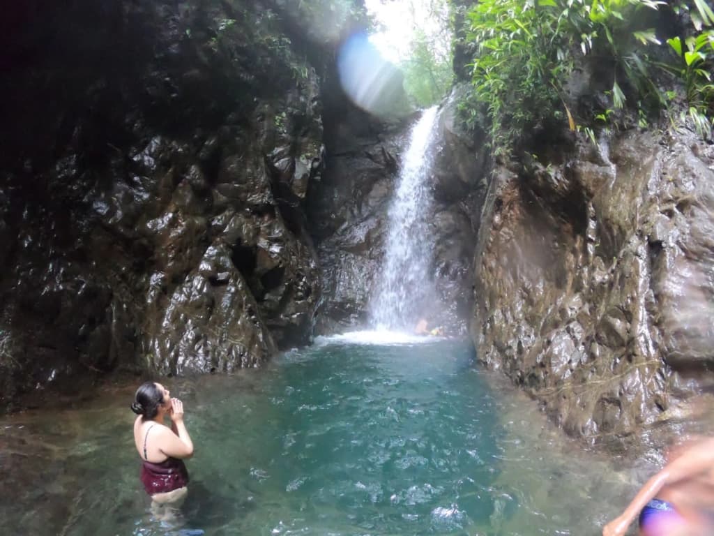 A woman stands near the edge of a natural pool, gazing at a waterfall surrounded by rocky terrain and lush greenery.