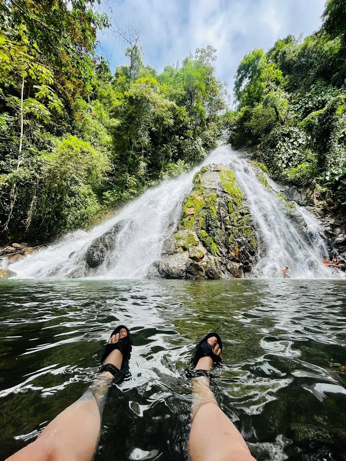 A person's legs in sandals are submerged in a clear pool of water at the base of a lush waterfall.
