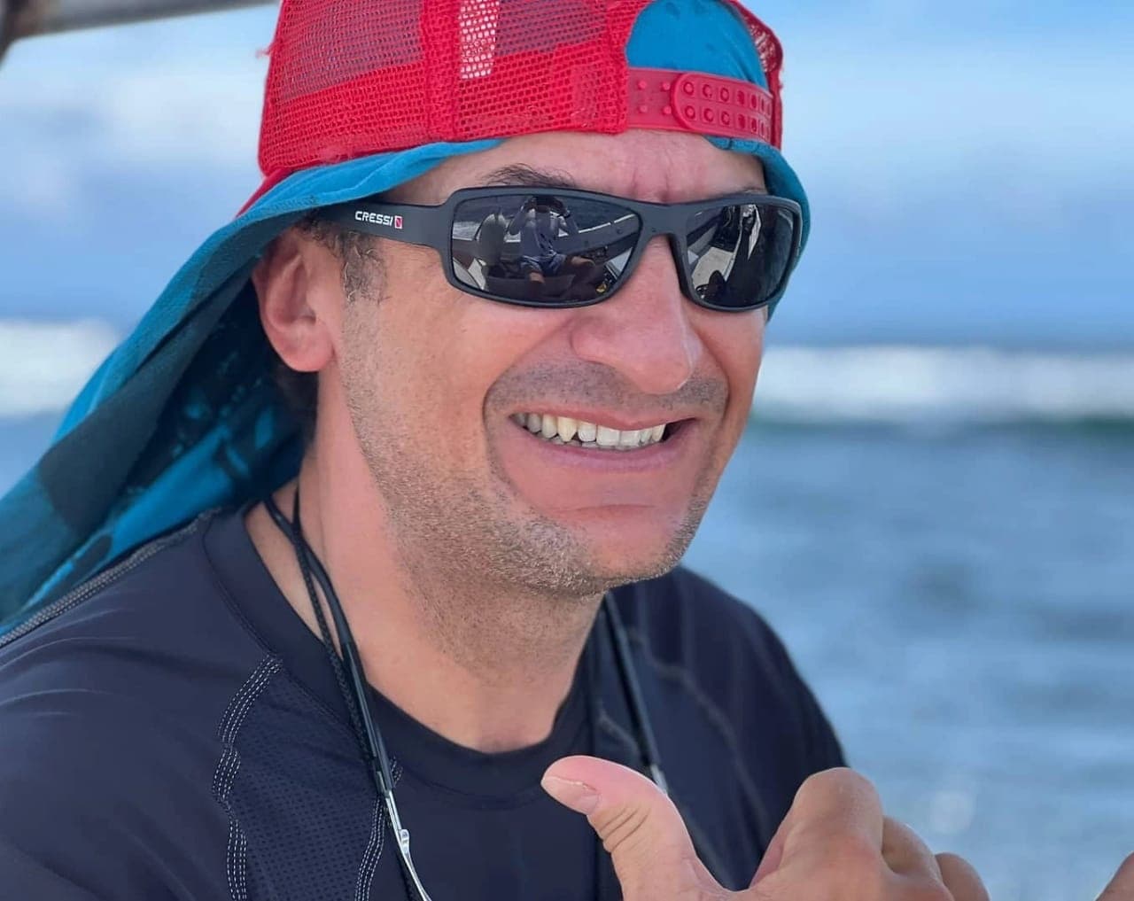 A smiling man wearing sunglasses and a colorful hat giving a thumbs-up near the water.