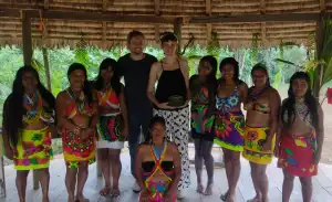 A group of people, including two individuals in casual attire, pose with several women dressed in colorful traditional clothing under a thatched roof.