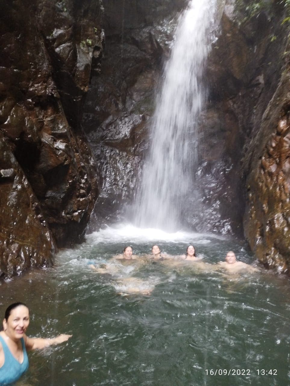 A group of five people enjoy swimming in a natural pool beneath a waterfall surrounded by rocky cliffs.