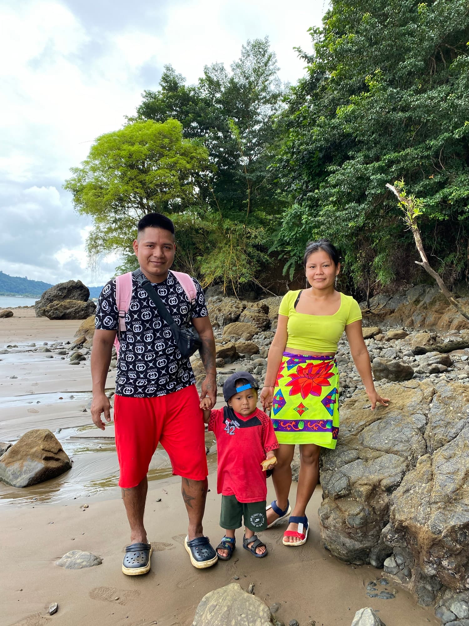 A family of three stands on a beach, surrounded by rocks and greenery.
